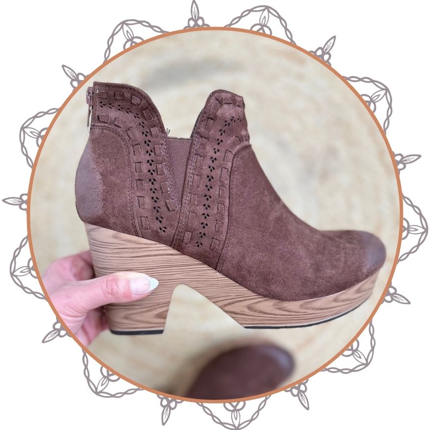 Brown suede boots on a wooden block with a hand holding one boot, on a decorative circular background.
